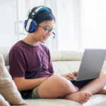 Boy sits on couch wearing headset playing game on laptop