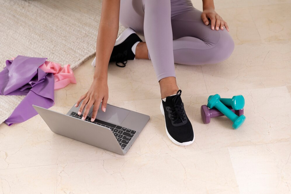 A female personal trainer working with her computer, having fitness equipment around - carpet, gums and dumbbells. Source: Freepik