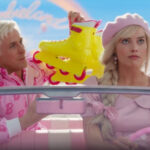Margot Robbie as Barbie, smiling and holding a microphone, on a pink stage with a background of cheering people