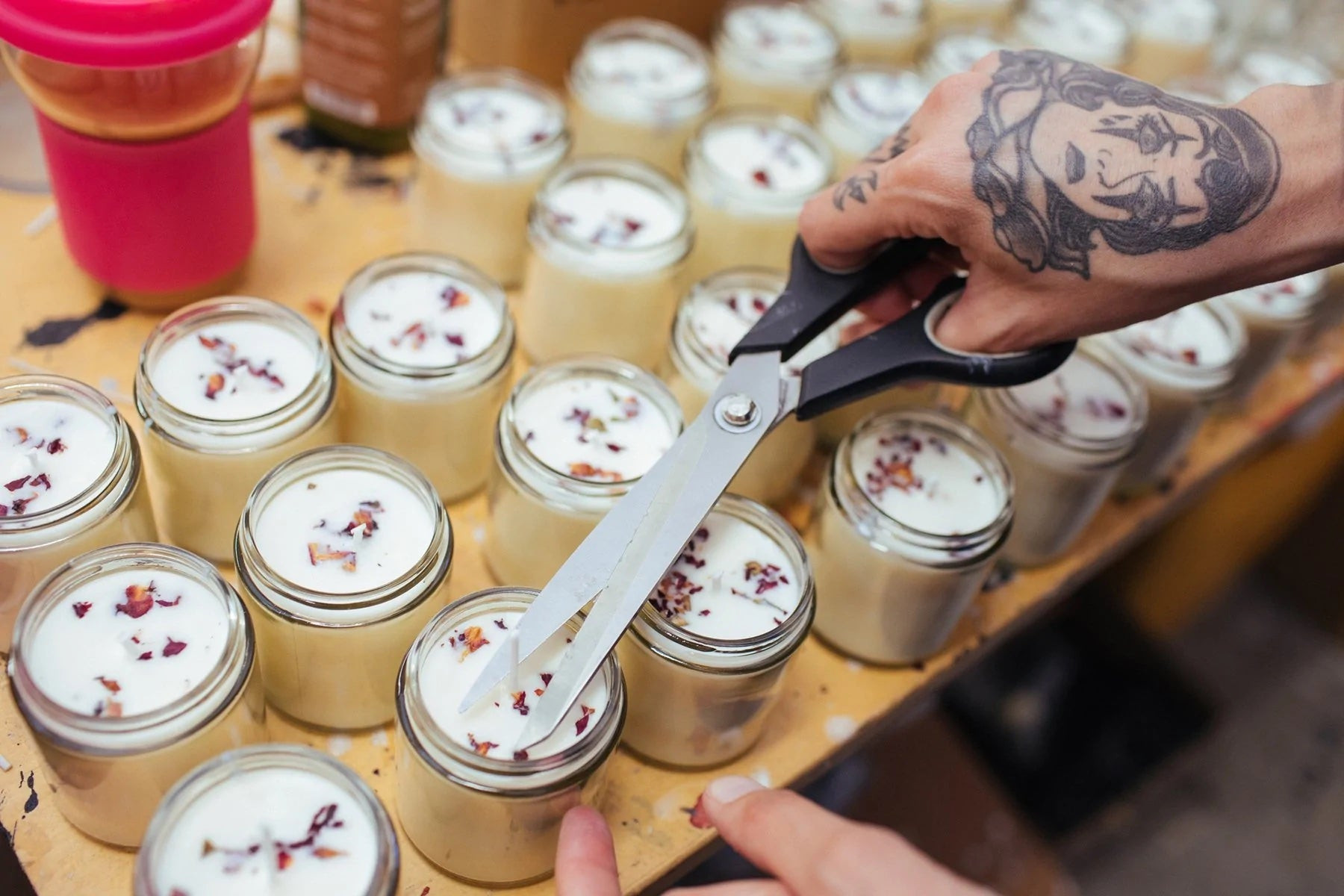A candle maker cuts wicks on a batch of candles arranged on a wooden table