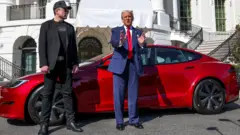 Donald Trump talks while standing next to Tesla CEO Elon Musk, with a red Tesla car and the White House in the background.
