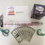 Supplies for creating a money box present, including a small box, wrapping paper, scissors, tape, a printable pull tab, and a utility knife